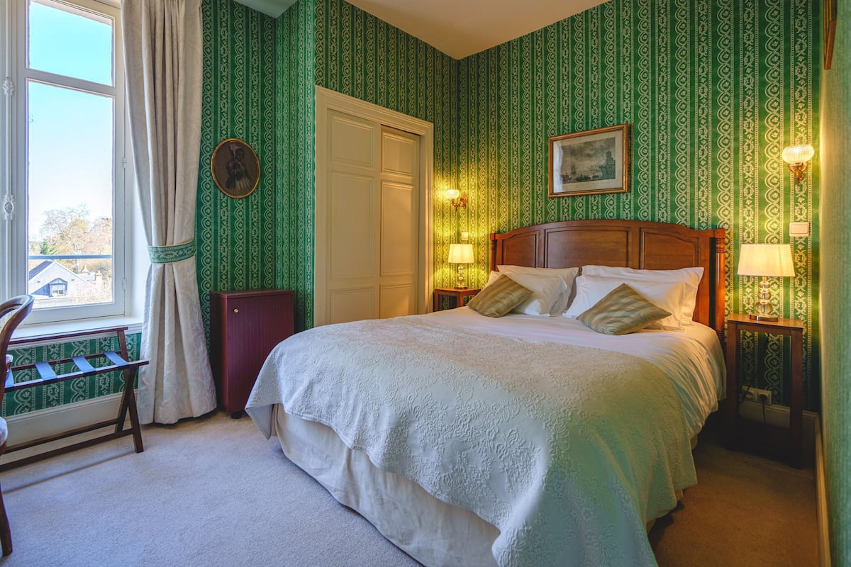 A bedroom with a large bed, a window with curtains, a desk, and a chair.