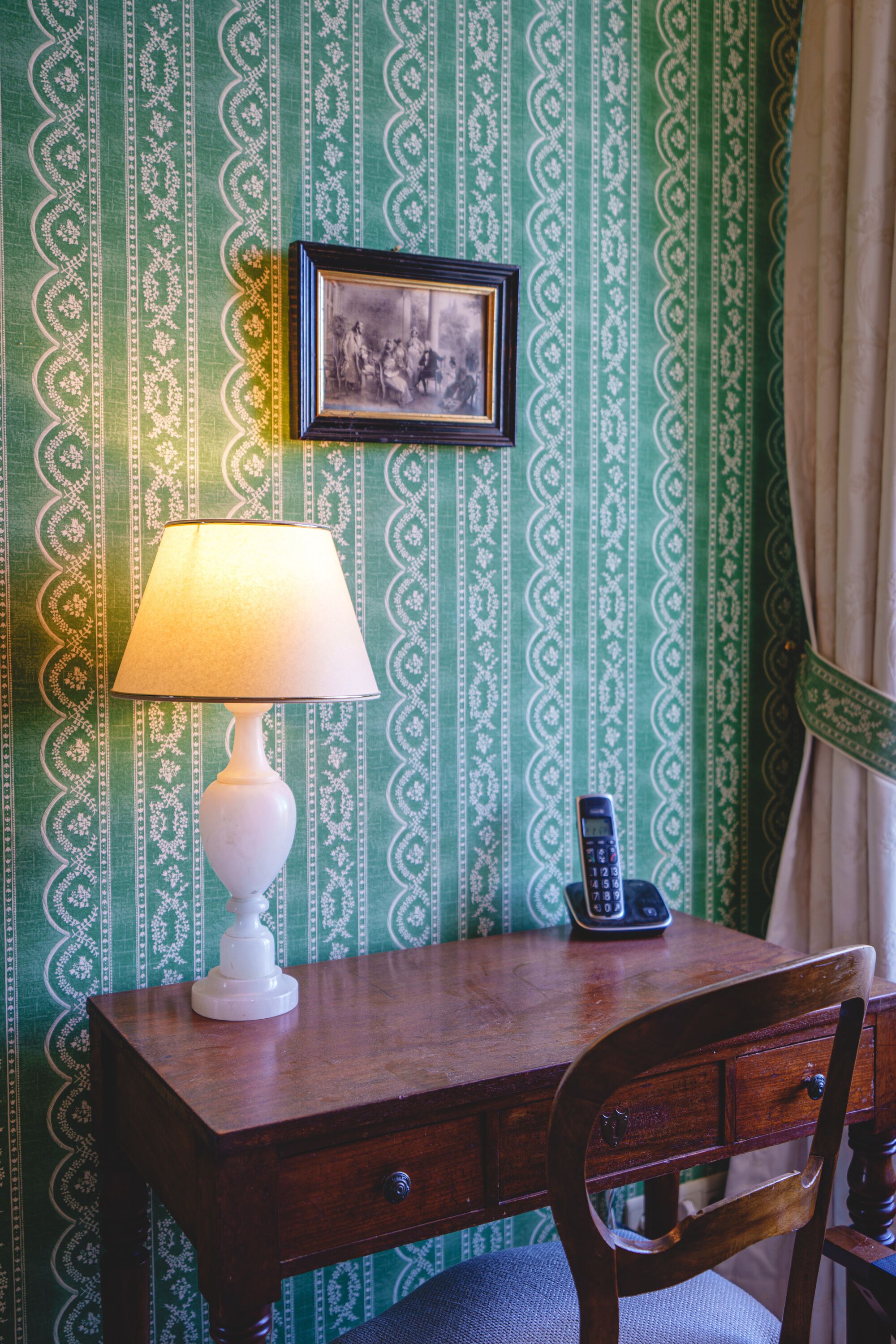 A wooden desk with a white lamp, a framed picture, and a telephone against a green patterned wallpaper.