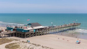 Eine Pier, die sich ins Meer erstreckt, mit einem Gebäude am Ende, einem Sandstrand und einem wolkenlosen Himmel.