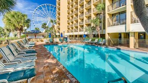 Ein Hotelpool mit Riesenrad im Hintergrund, Liegestühlen und einem Gebäude im tropischen Stil.