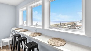A room with a long counter, four black stools, and three large windows with starfish decorations. - Waves of Joy: Oceanfront Home with Elevated Pool and Elevator (Oak Island)