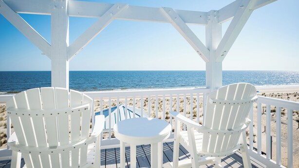 A beachside deck with white Adirondack chairs and a small table, overlooking the ocean. - Waves of Joy: Oceanfront Home with Elevated Pool and Elevator (Oak Island)
