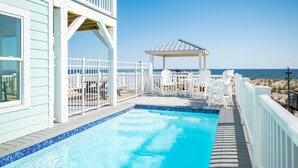 A pool area with a clear view of the ocean, a covered seating area, and a white fence. - Waves of Joy: Oceanfront Home with Elevated Pool and Elevator (Oak Island)