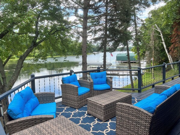 Une terrasse dotée de meubles en osier et de coussins bleus, offrant une vue sur un lac où naviguent des bateaux et située à proximité d’une zone boisée.