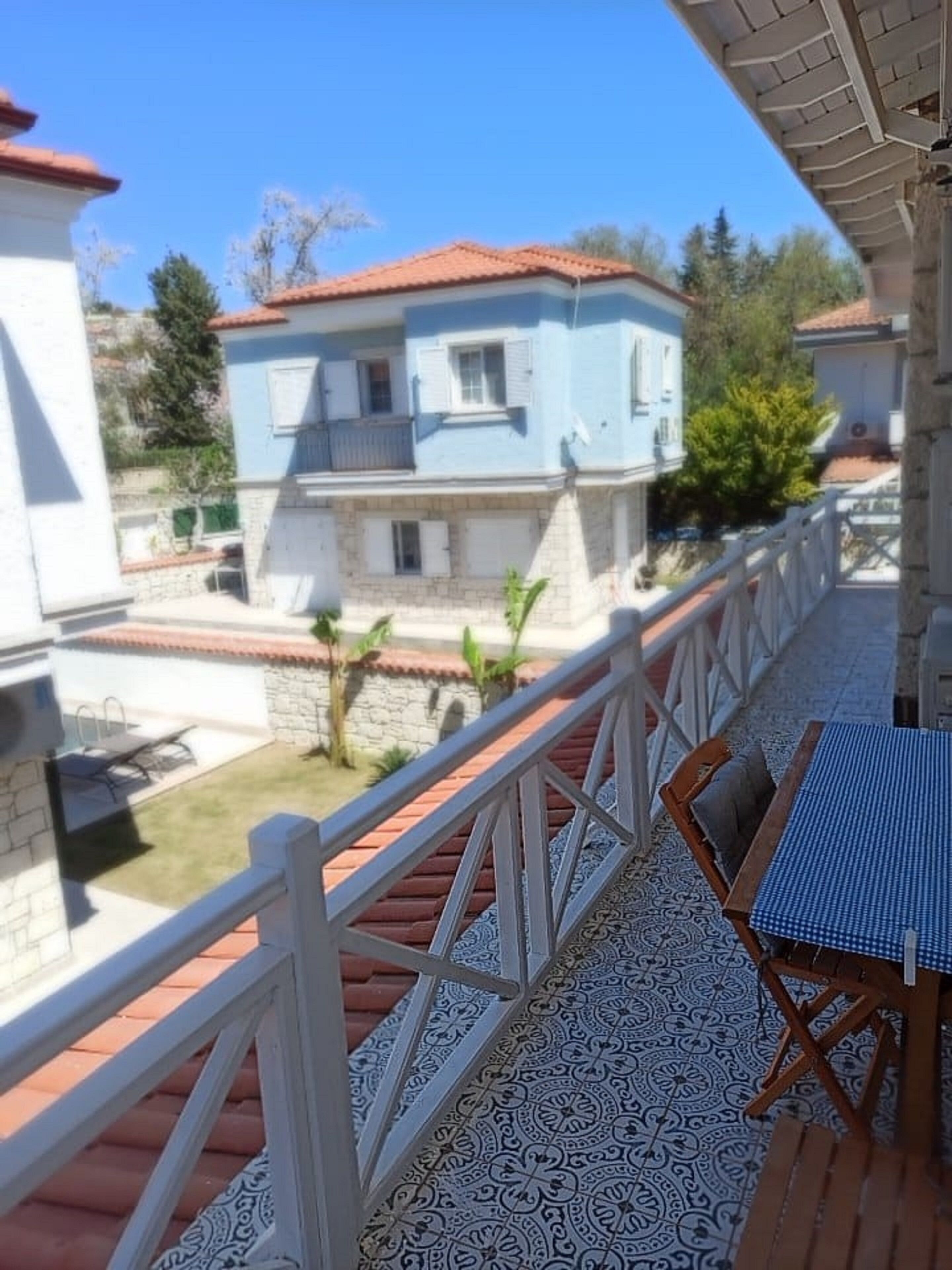A balcony with a patterned floor, a table with chairs, and a view of a two-story building with a red-tiled roof.
