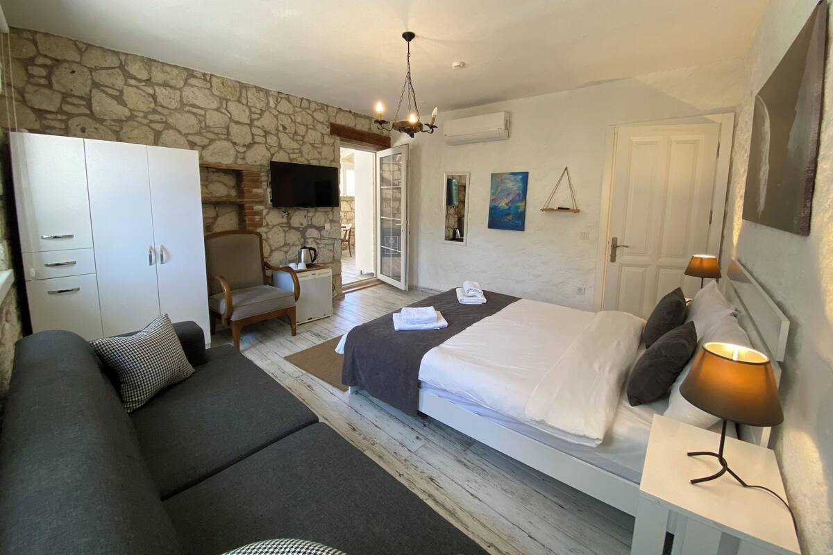 A bedroom with a bed, a sofa, a TV, a stone wall, and a door.