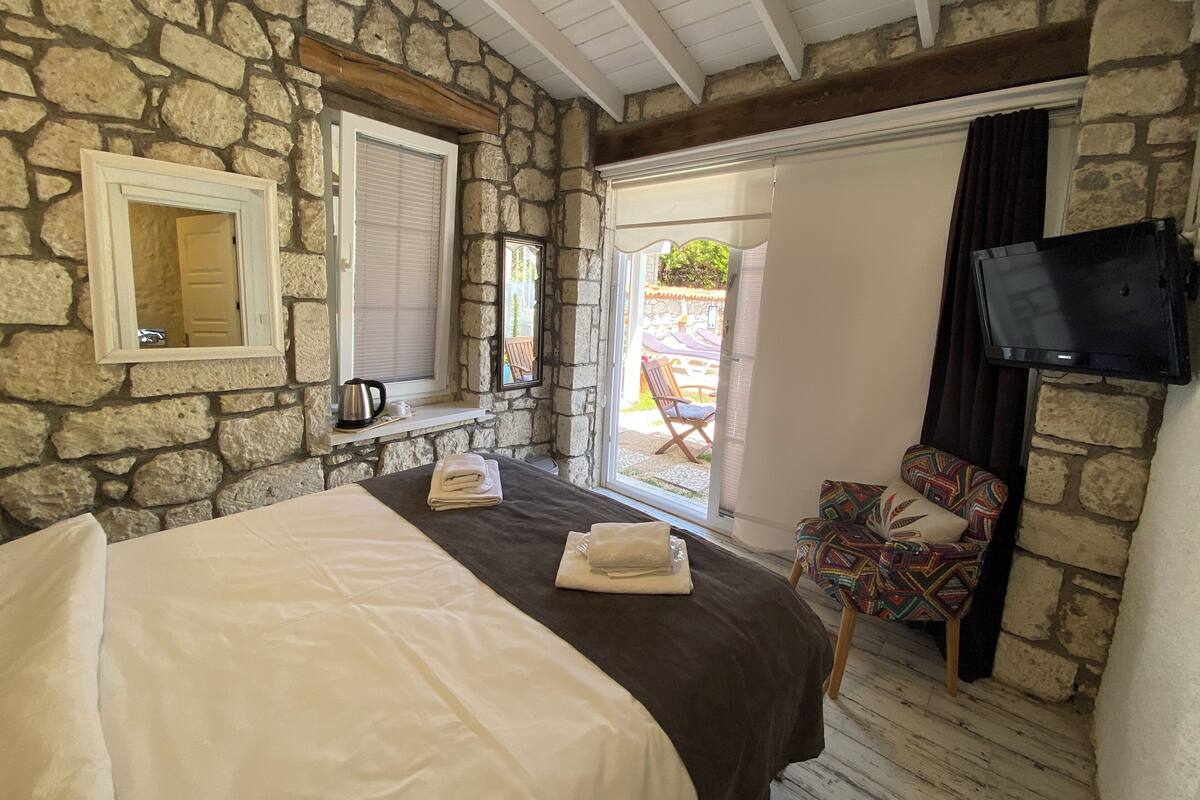 A bedroom with stone walls, a bed with white and dark bedding, a window with a view, a chair, and a television.