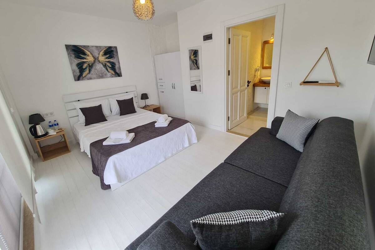 A bedroom with a bed, a grey sofa, a bedside table with a lamp, a wall-mounted artwork, and a door leading to a bathroom.