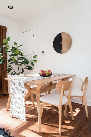 A dining area with a wooden table, white chairs, a round wall clock, and a potted plant.