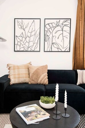 A modern living room with a black sofa, two framed abstract plant artworks, a round coffee table with a magazine, a small plant, and two candles.