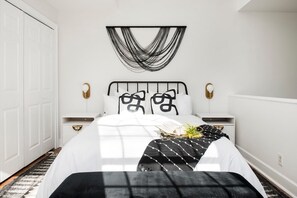 A bedroom with a bed, bedside tables, a wall-mounted light fixture, and a decorative wall hanging. - Checkmate (Atlanta)