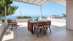 Un área de comedor al aire libre techada con vistas al mar, amueblada con sillas y una mesa de madera, rodeada de paredes blancas.