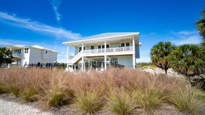 A two-story beachfront house with a balcony, surrounded by beach grass and palm trees. - Gulf Views, 4BR/4BA w/ Game Room, Private Pool w/ Heat, EV Charger (St. George Island)