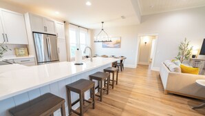 A modern kitchen with a large island, wooden stools, and a dining area with a sofa and table. - Gulf Views, 4BR/4BA w/ Game Room, Private Pool w/ Heat, EV Charger (St. George Island)