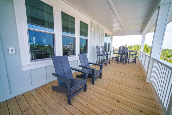 A covered porch with wooden flooring, two Adirondack chairs, a small table, and a railing. - Gulf Views, 4BR/4BA w/ Game Room, Private Pool w/ Heat, EV Charger (St. George Island)