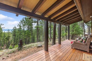A wooden deck with a wicker sofa and a red throw pillow, overlooking a forested area.