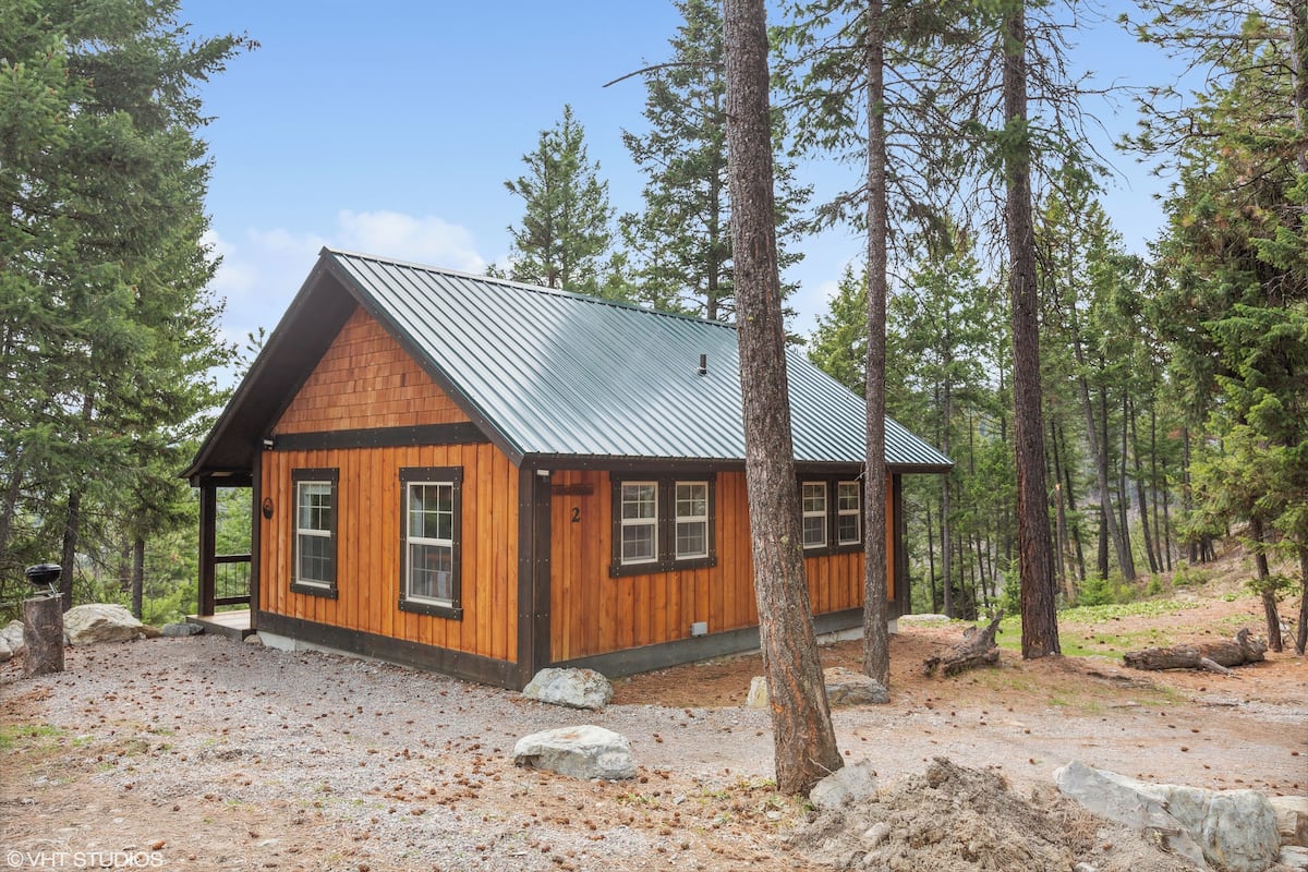 A cabin with a sloped metal roof, wooden walls, and a gravel driveway surrounded by trees.