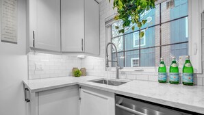 A modern kitchen with white cabinets, a stainless steel dishwasher, and a window with a view of a blue building. - A bright & chic 2 bedroom urban apartment (Orangeville)
