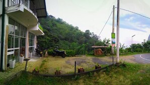 A building with large windows, a paved area with potted plants, and a road curving to the right. - Dewy Arena Hotel (Ella)