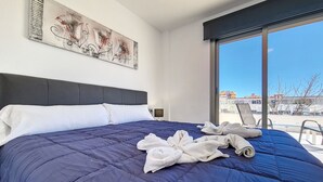 Cama bem arrumada com edredom azul escuro e almofadas brancas, uma grande pintura abstrata na parede e vista de prédios por uma janela ampla.