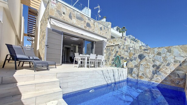 Uma área de piscina moderna ao ar livre, com espreguiçadeiras, conjunto de jantar e parede de pedra.