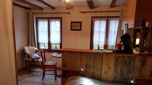 Um quarto acolhedor com uma cama, uma escrivaninha de madeira, uma cadeira e uma mesa. Há janelas com cortinas, um quadro na parede e uma cafeteira na escrivaninha.