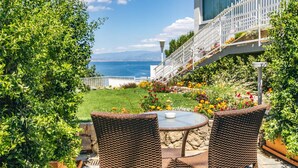 Eine Terrasse mit Korbstühlen und einem Glastisch, mit Blick auf einen Garten voller bunter Blumen und das Meer.