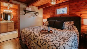 Een gezellige slaapkamer met een houten plafond, een groot bed met beddengoed met een patroon, een nachtkastje met een fles en glazen, en een plafondventilator.