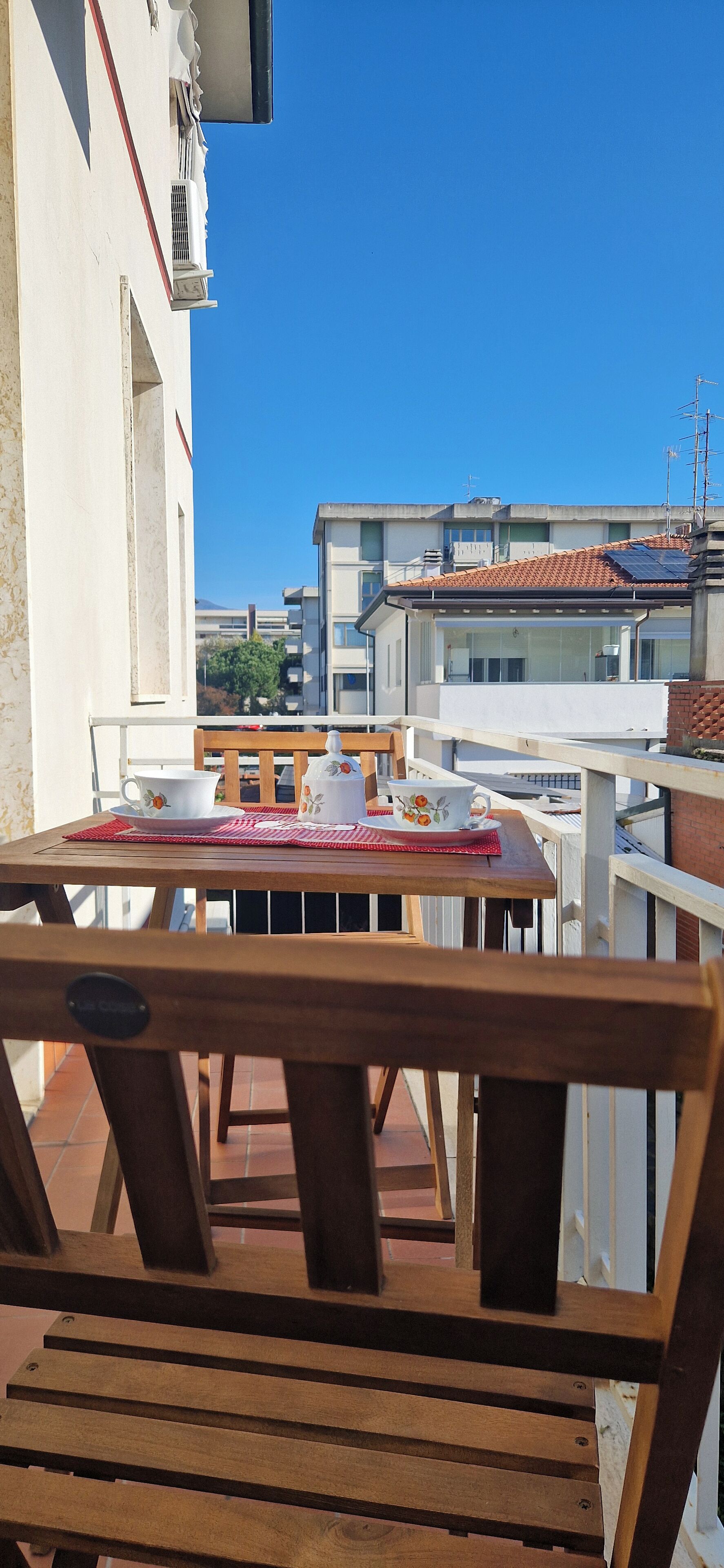 Ein Holztisch mit einer roten Tischmatte und zwei weißen Tassen mit floralen Mustern auf einem Balkon.
