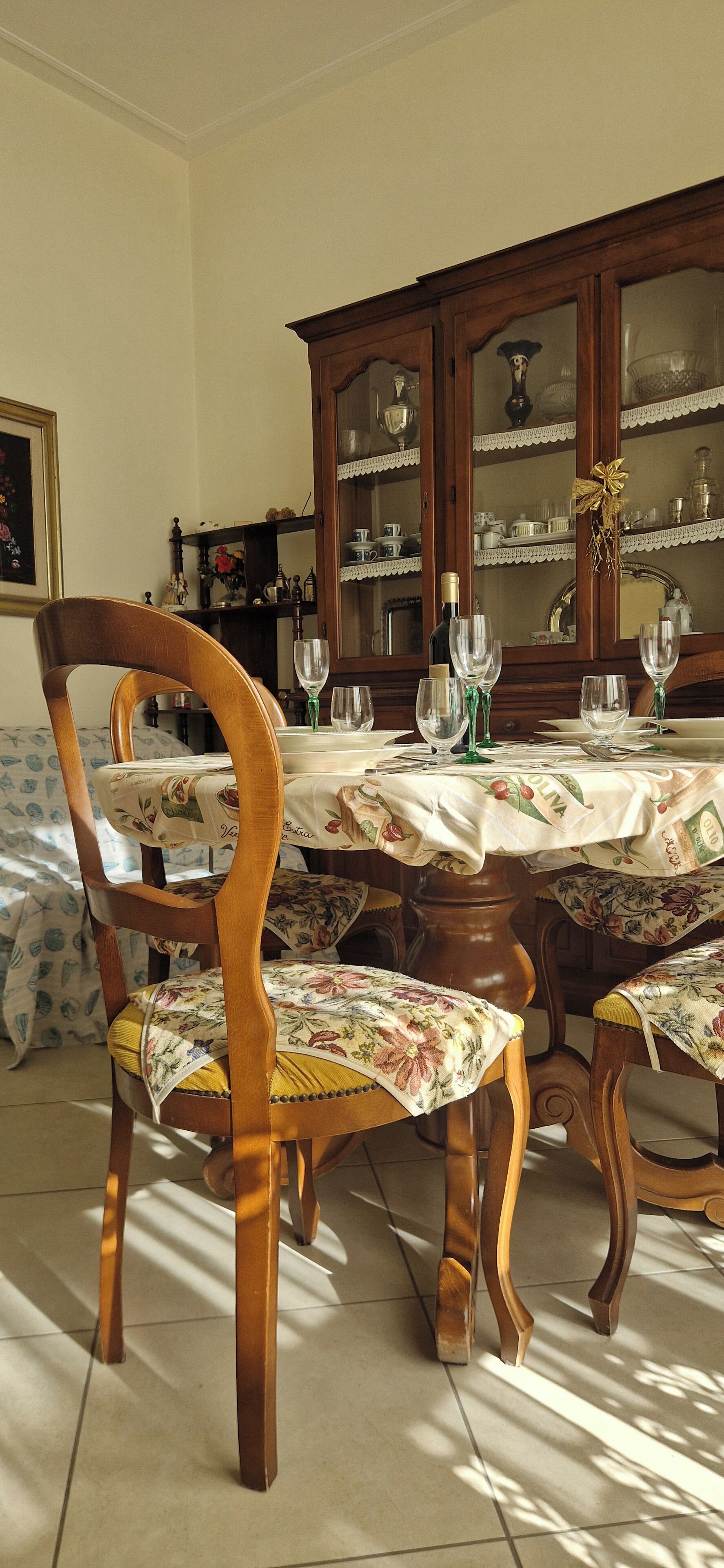 A dining room with a wooden table set for four, surrounded by chairs with floral upholstery. A wooden cabinet with glass doors is in the background, displaying various items.