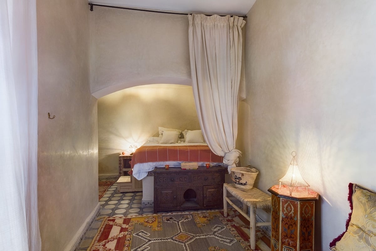A narrow hallway leading to a bedroom with a bed, a nightstand, a lamp, and a patterned rug.