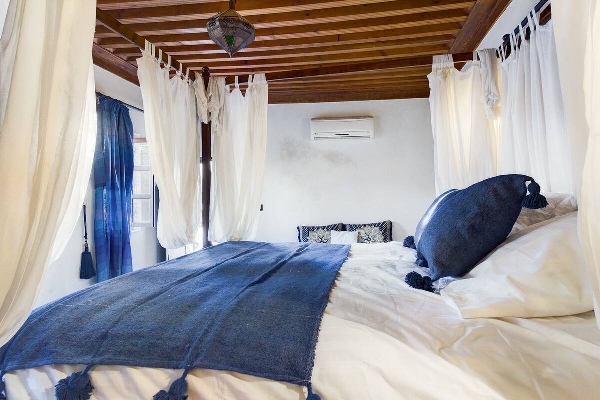 A bedroom with a wooden ceiling, white curtains, a bed with a blue blanket, and a hanging lantern.