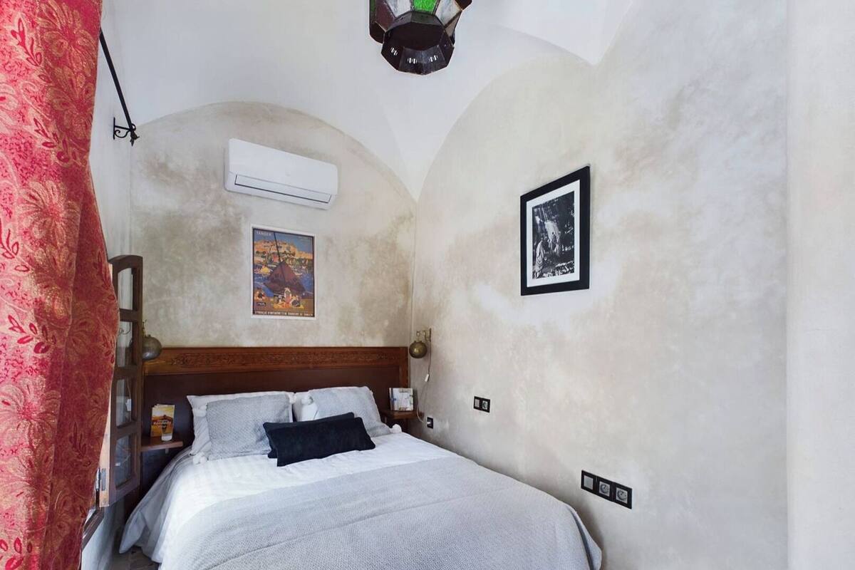 A bedroom with a wooden headboard, a bed with a white and gray blanket, an air conditioning unit, a framed picture, and a red curtain.