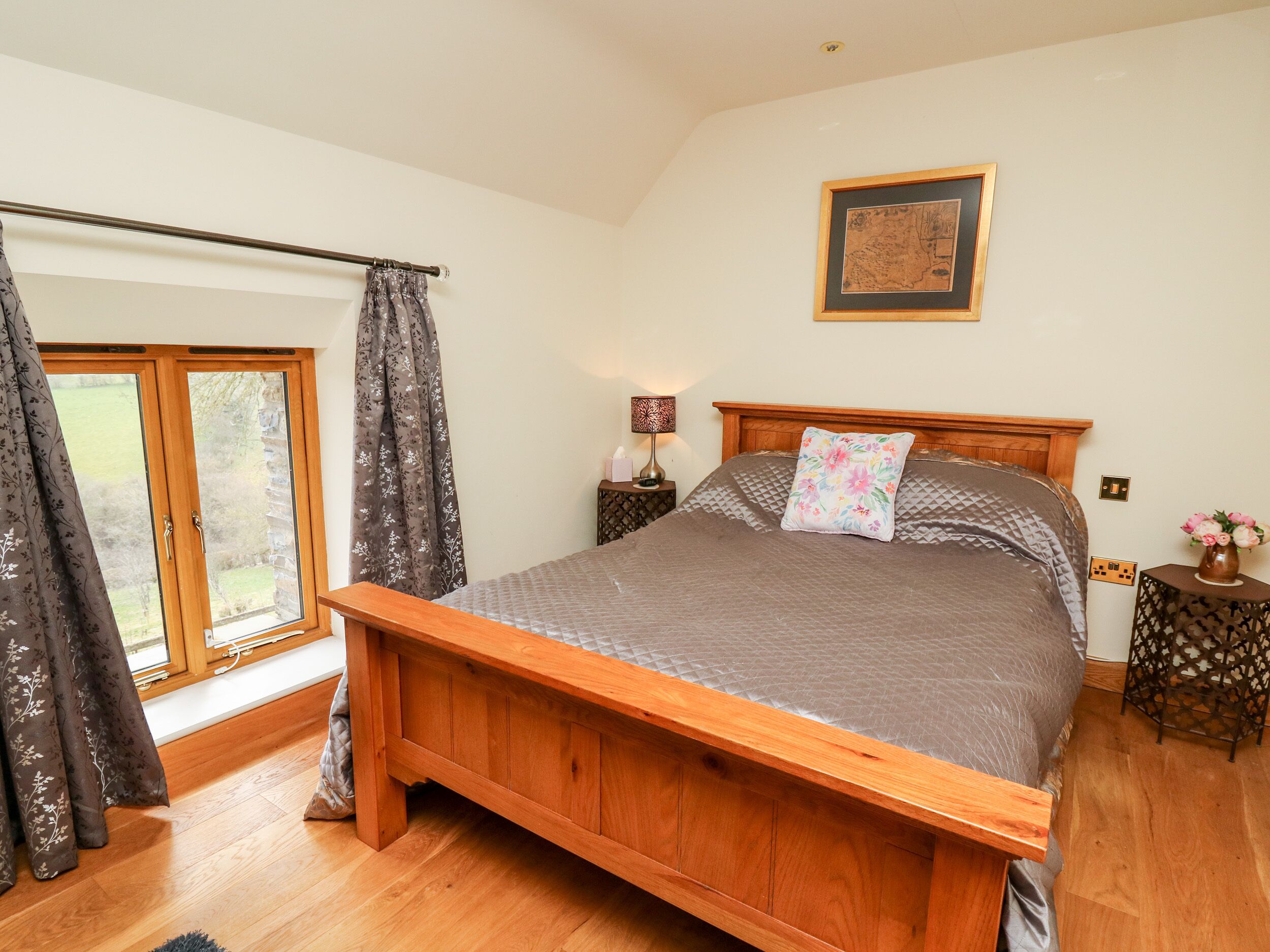 A bedroom with a wooden bed, a window with curtains, a bedside table with a lamp, and a framed picture on the wall.