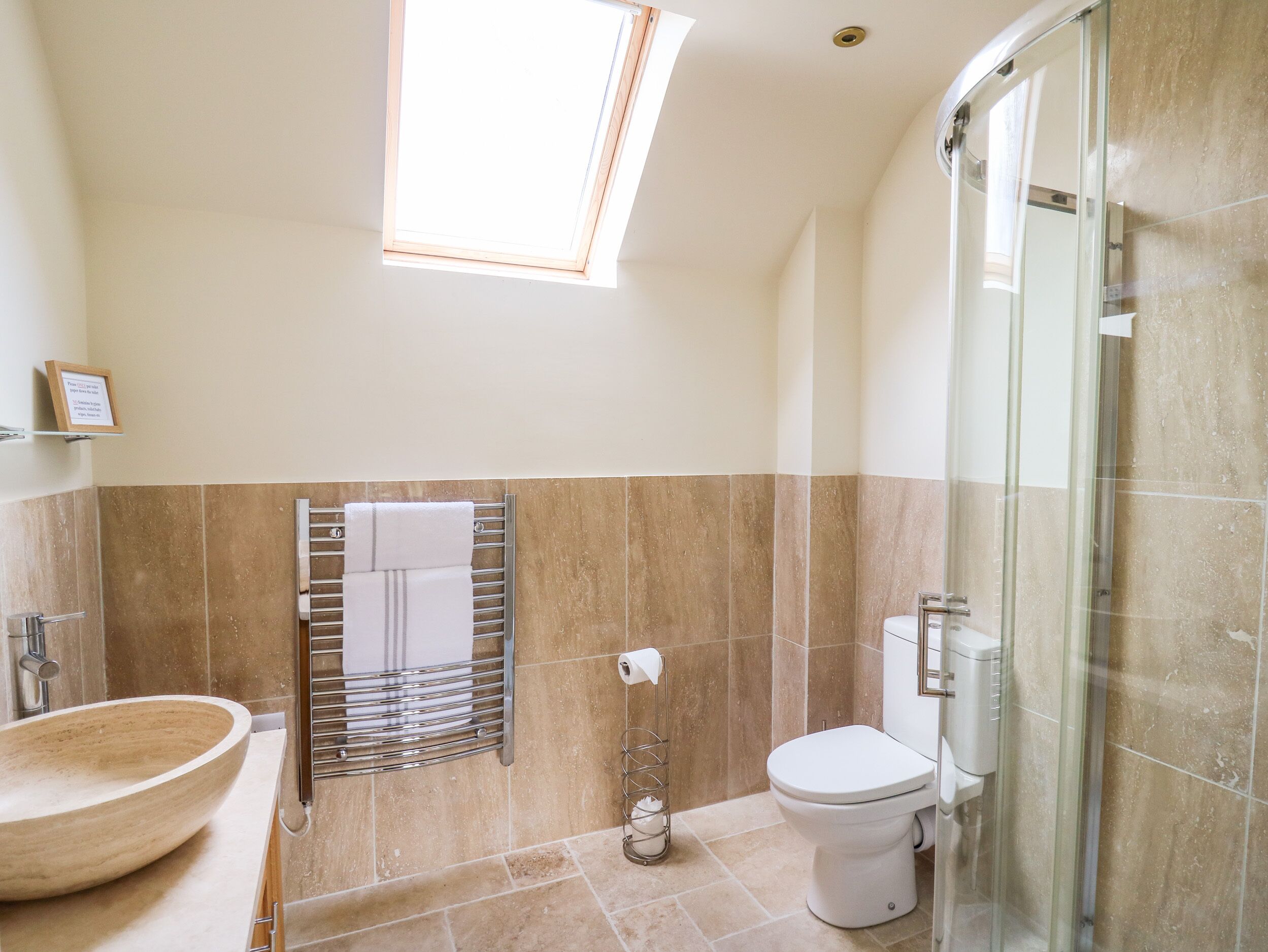 A bathroom with a skylight, a wooden basin, a towel rack, a toilet, and a glass shower enclosure.