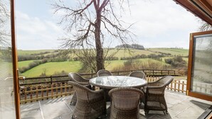 A covered outdoor patio with wicker furniture and a glass table, overlooking a rural landscape. - The Orchard (Knighton)
