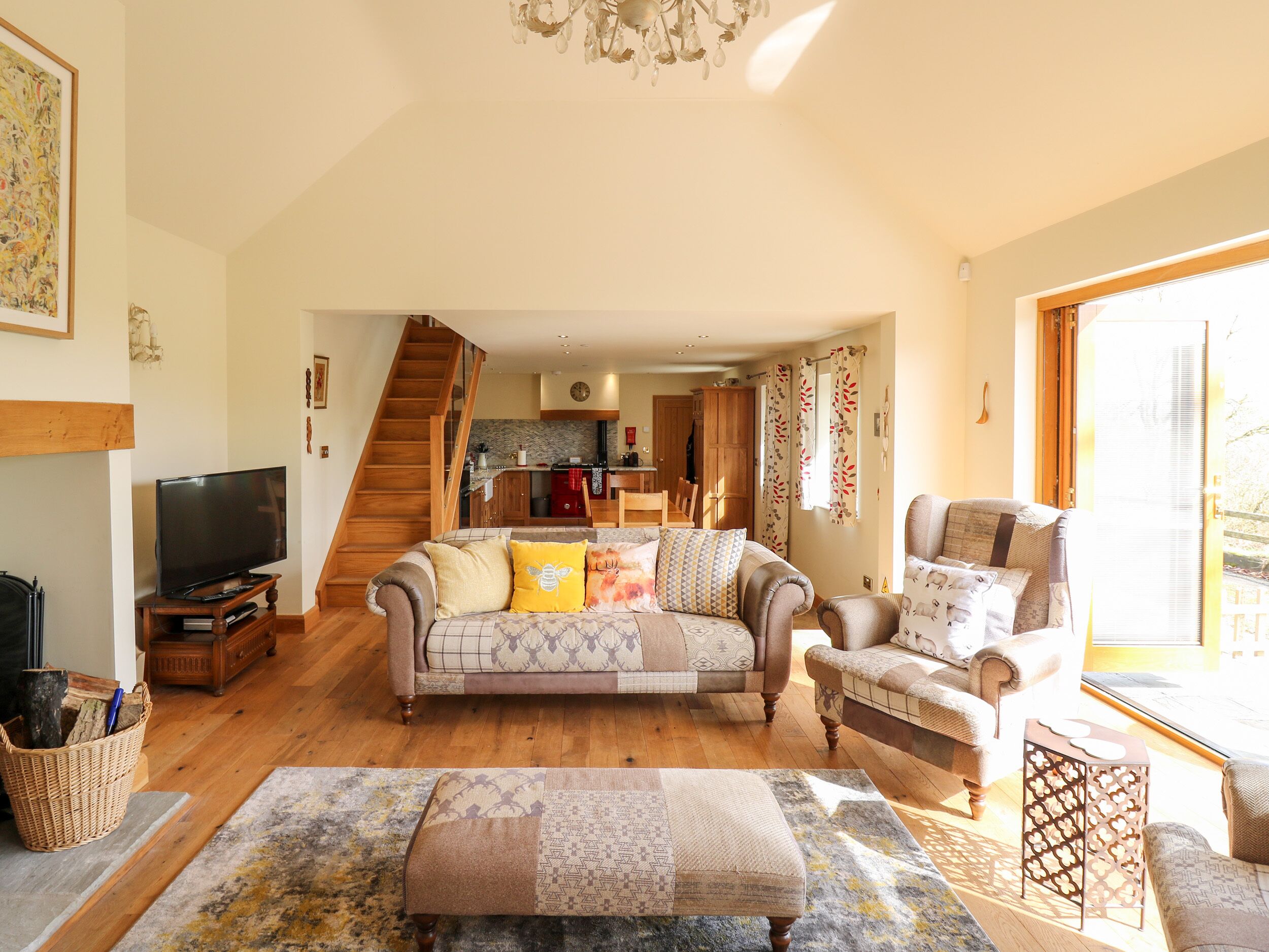 A spacious living room with a sofa, armchairs, a TV, and a staircase leading to a kitchen.