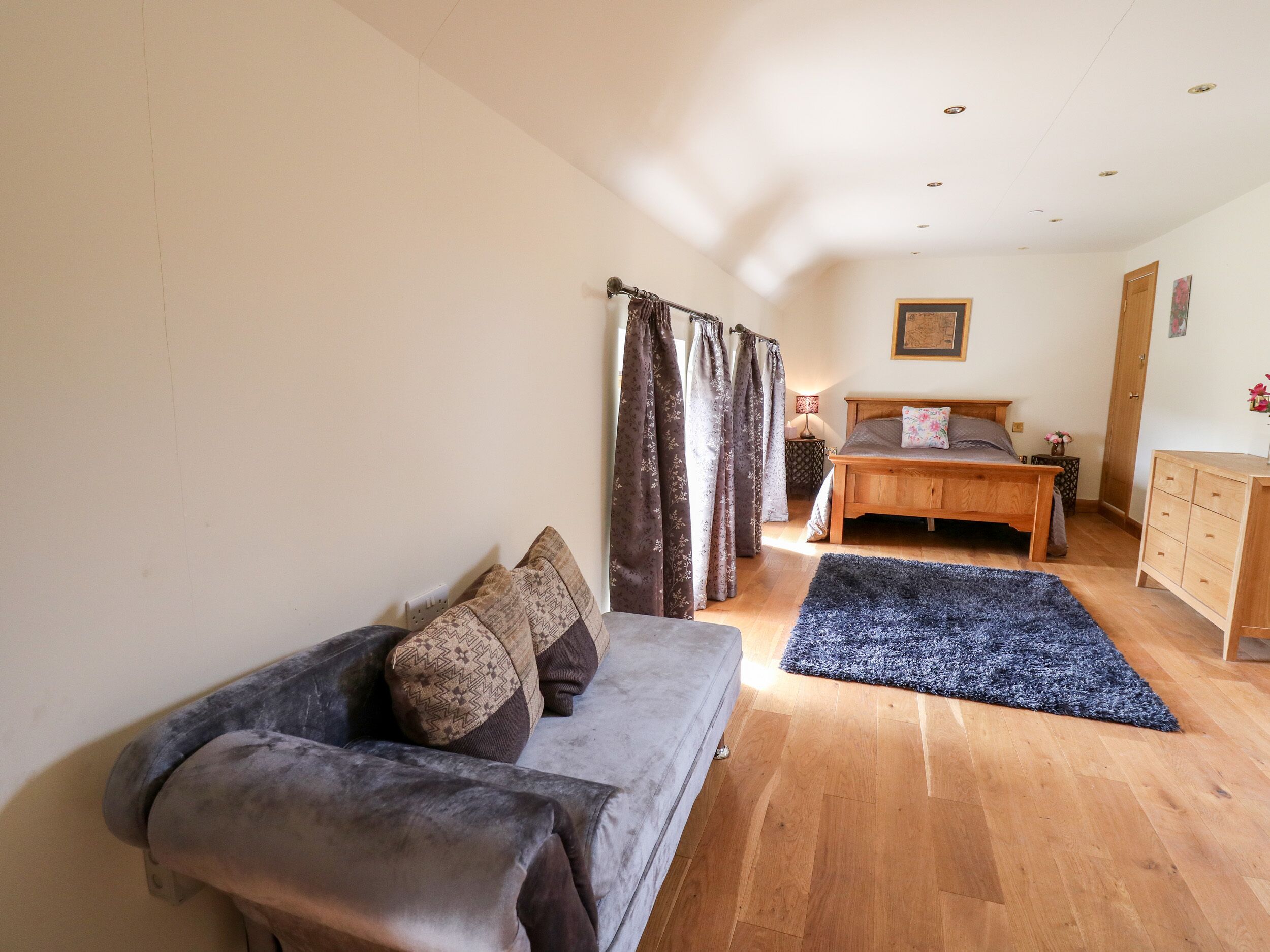 A bedroom with a wooden bed, a grey sofa, a blue rug, and a wooden dresser.