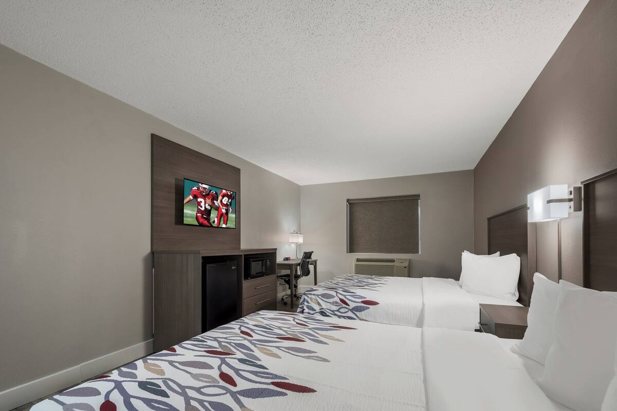 A hotel room with two beds, a desk with a chair, a TV mounted on the wall, and a window.