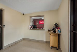 A hotel corridor with a mirror reflecting a red sign, a small cart with cleaning supplies, and a door on the left.