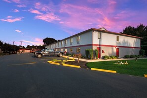 Un bâtiment avec une porte et des fenêtres rouges, un parking avec un véhicule noir, et une zone herbeuse avec de petites plantes.
