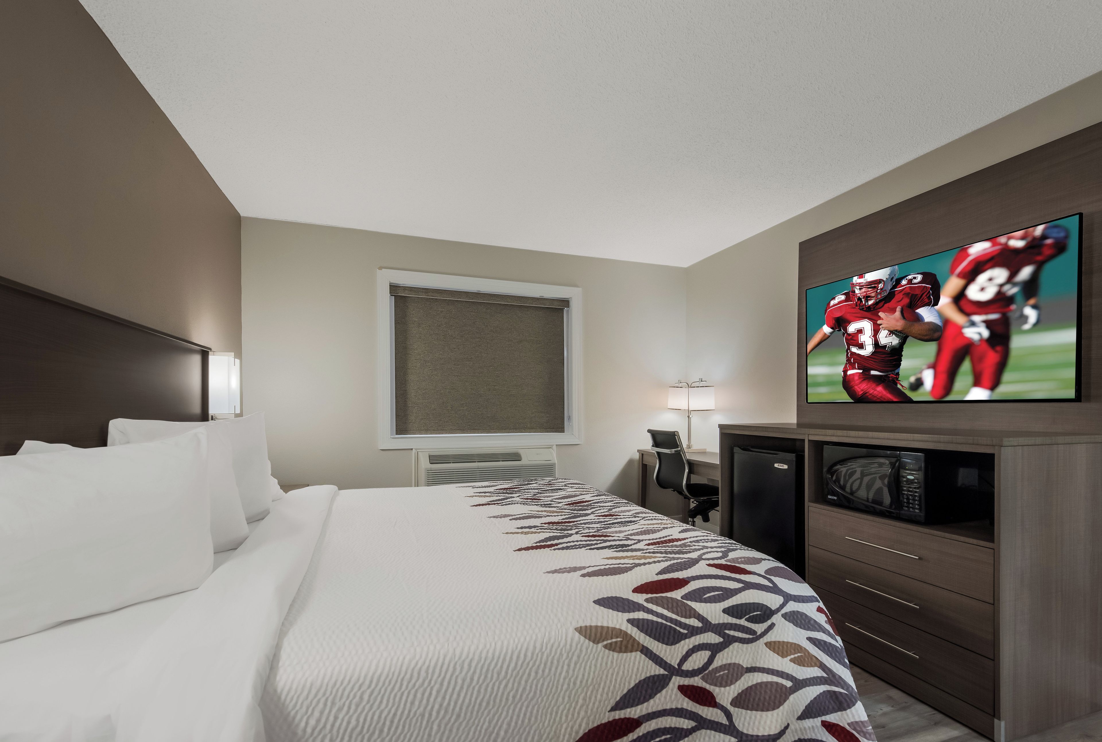 Quarto de hotel com uma cama grande, televisão fixada na parede, escrivaninha com cadeira e micro-ondas no canto.