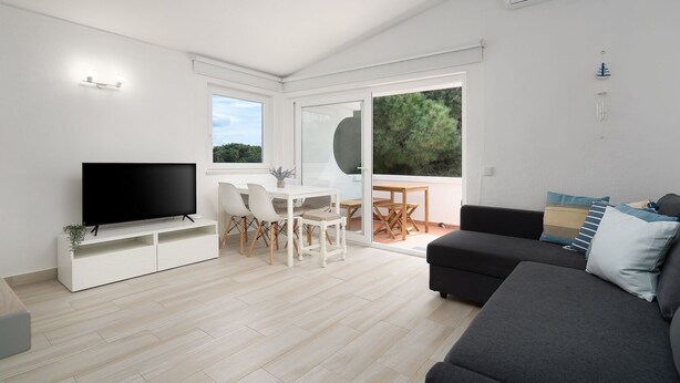 Ein modernes Wohnzimmer mit Flachbildfernseher, Essbereich mit Tisch und Stühlen, einer Couch und einem großen Fenster mit Blick ins Grüne.