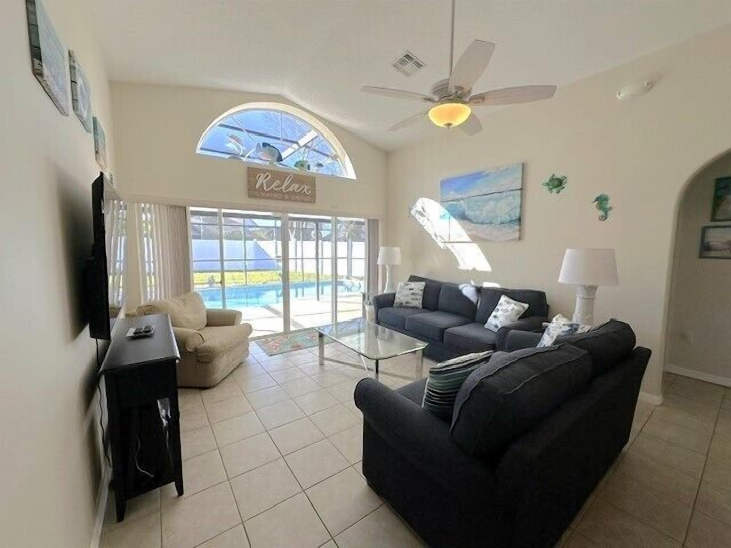 A living room with a sofa, a coffee table, a TV, and a ceiling fan. There is a large window with a view of a pool and a sign that says "Relax".