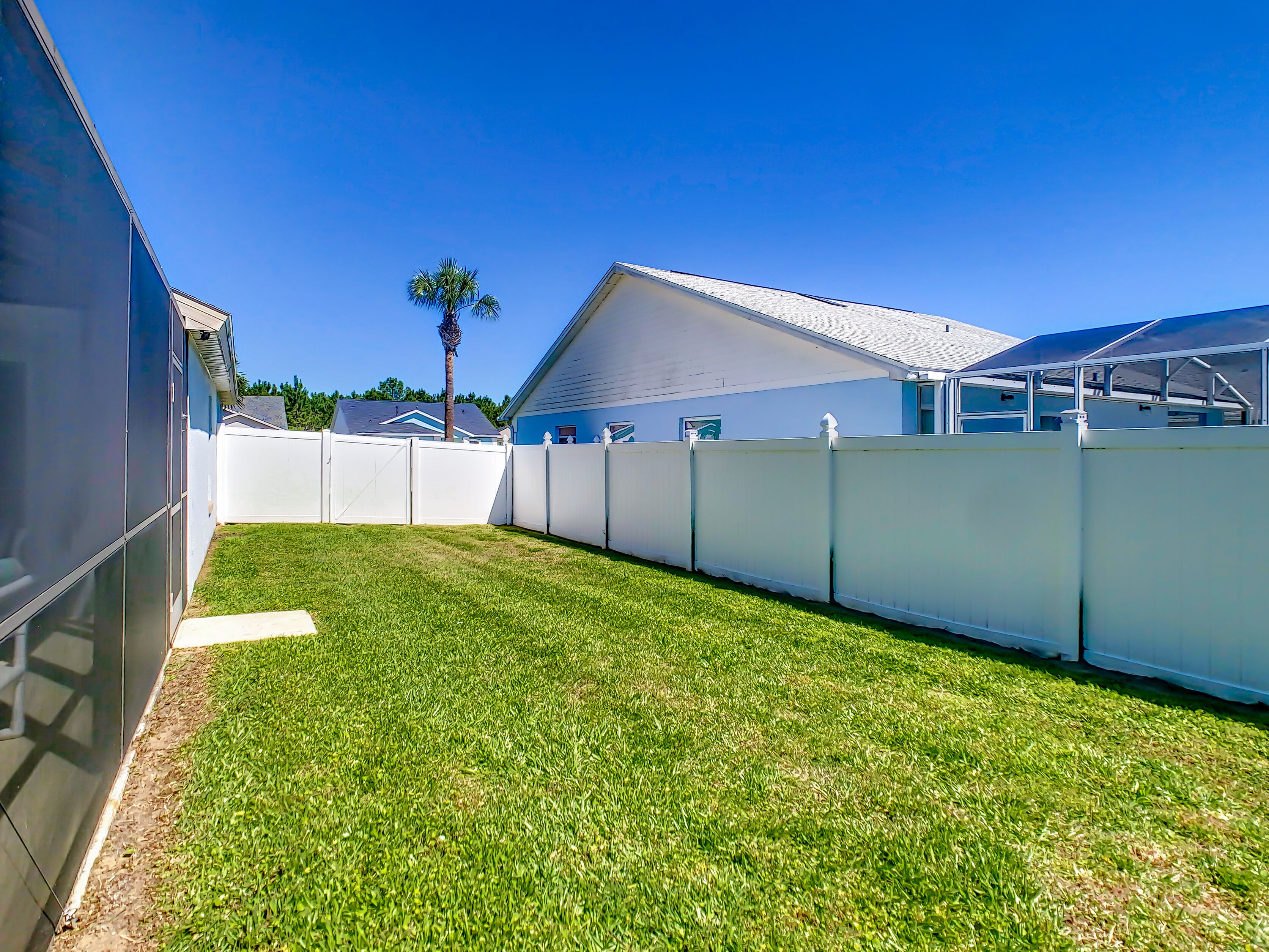 Un patio trasero residencial con una cerca blanca, un césped y una casa con techo inclinado.