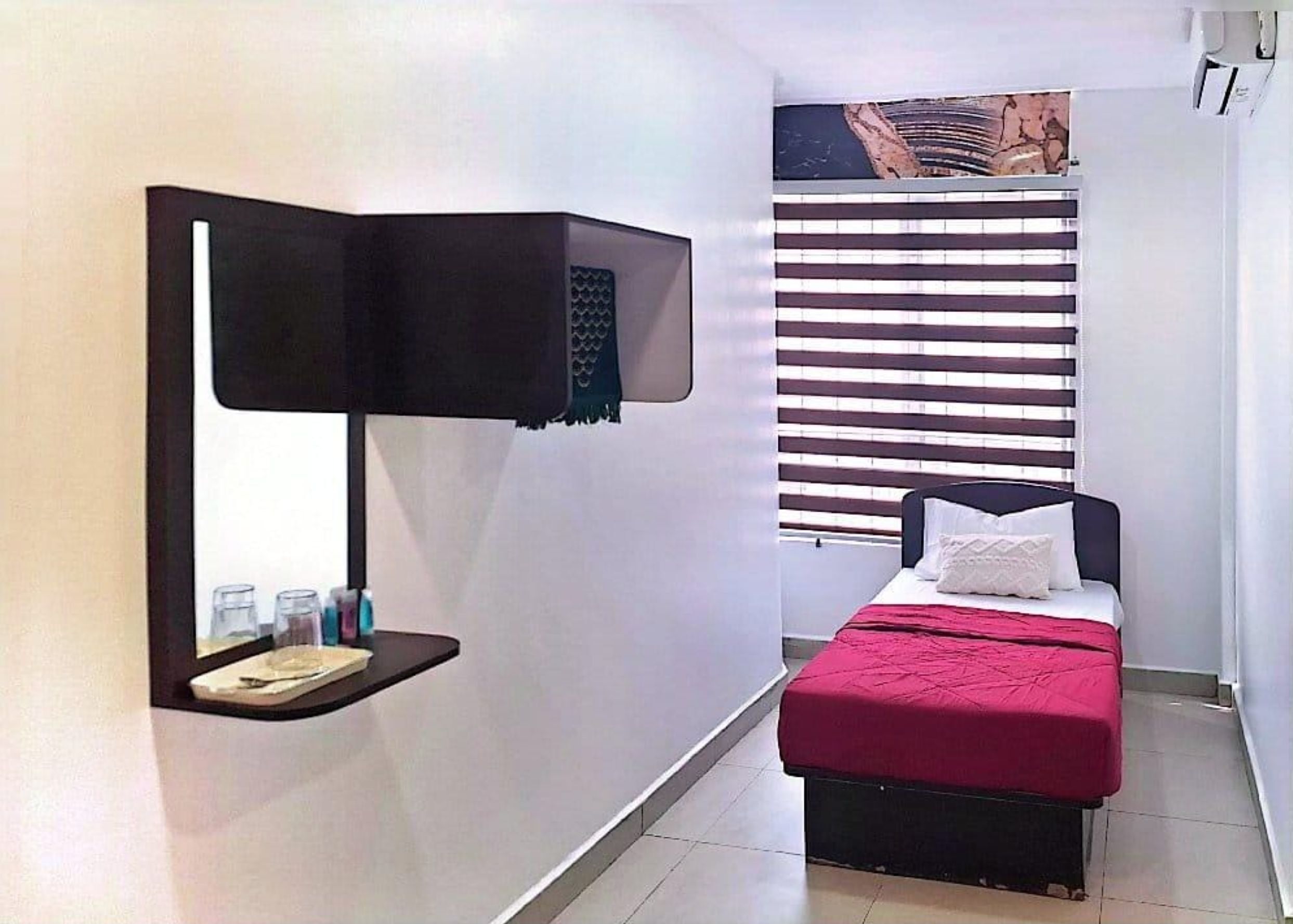 A small, modern bedroom with a bed, a bedside table, a wall-mounted lamp, and a window with blinds.