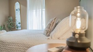 Ein Schlafzimmer mit einem Bett, einem Nachttisch mit Lampe, einem Spiegel und einem Fenster mit Vorhängen.