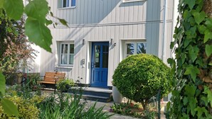 Un edificio de dos pisos con puerta y ventanas azules, rodeado de vegetación y un banco.
