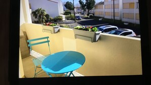 Ein Balkon mit blauem Stuhl und Tisch, Topfpflanzen und Blick auf eine Straße mit parkenden Autos und Gebäuden.