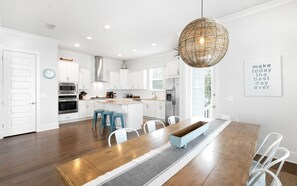 A modern kitchen with white cabinetry, a central island, and a dining area with a wooden table and blue stools. - Inlet Beach Home + Pool, Pet Friendly & Bikes (Inlet Beach)